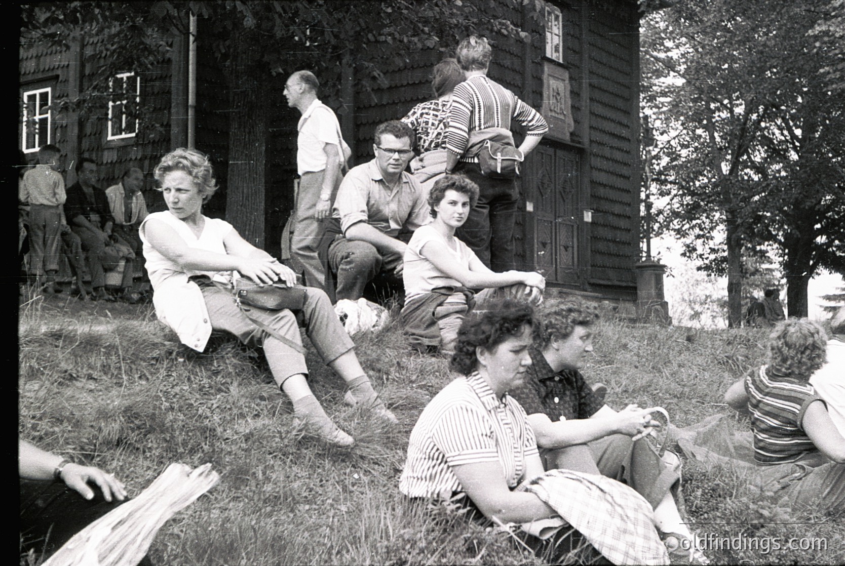 Group of young people lounging on grass near a rustic wooden house, mid-20th century. Casual summer attire—shorts, striped shirts, and sandals—suggests a relaxed, informal gathering. Wooden shutters and overgrown foliage hint at rural or suburban setting.