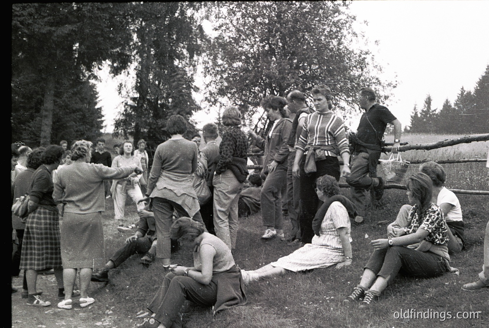 Vintage outdoor gathering of young adults in 1960s-70s attire, likely a youth camp or social event. Group of ~20 people in casual summer clothes—plaid skirts, button-ups, and headscarves—engaged in conversation and activities near a wooden fence and trees. One person sits on a blanket, others lean against the fence or stand in a circle. Rural or park setting with natural lighting.