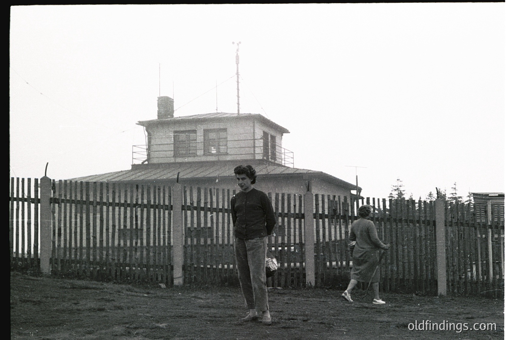 1960s-era watchtower with cylindrical observation deck, surrounded by a chain-link fence. Two individuals in casual mid-century attire—one holding a ball—pose in front. Likely a coastal or border surveillance post.