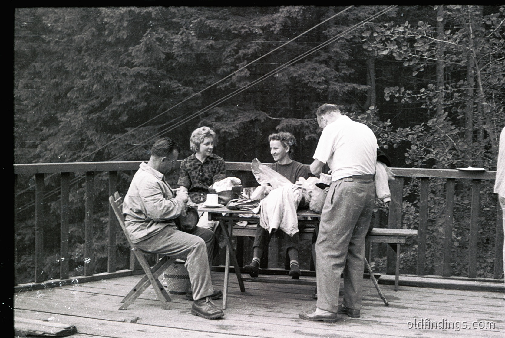 Mid-20th century group picnic on a wooden deck overlooking forested hills. Four adults seated/standing around a small table, dressed in 1950s-60s attire—men in short-sleeve button-ups and trousers, women in dresses. One man holds a cup, another reads a newspaper. Vintage black-and-white film captures relaxed outdoor socializing.