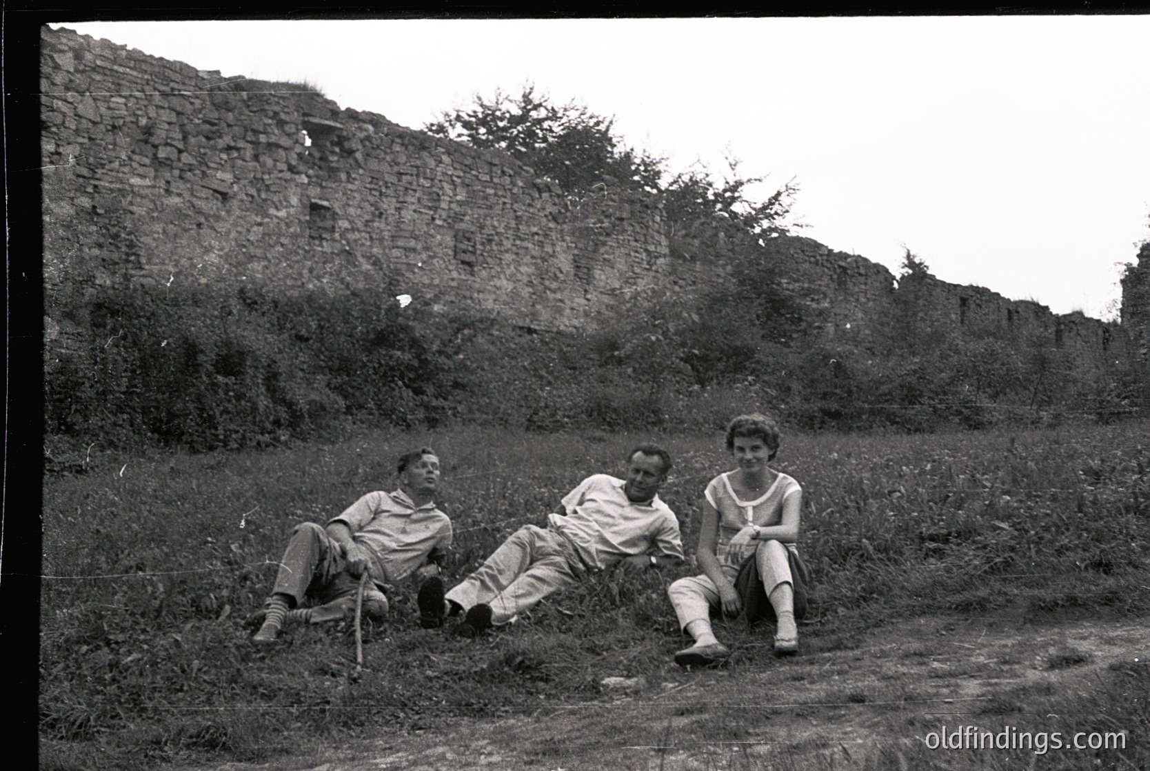Three individuals pose casually in mid-20th-century attire—lightweight shirts, trousers, and sandals—against a weathered stone wall and overgrown grass. Likely or outdoor portrait.
