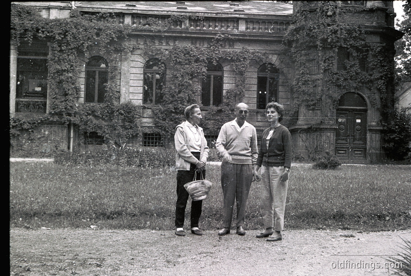 Three individuals pose outdoors in front of a grand, ivy-covered building with arched windows and ornate stonework, likely mid-20th century. The man in the center wears a sweater and trousers; the woman on the left holds a woven basket. The scene suggests a formal or institutional setting, possibly a university or cultural center.
