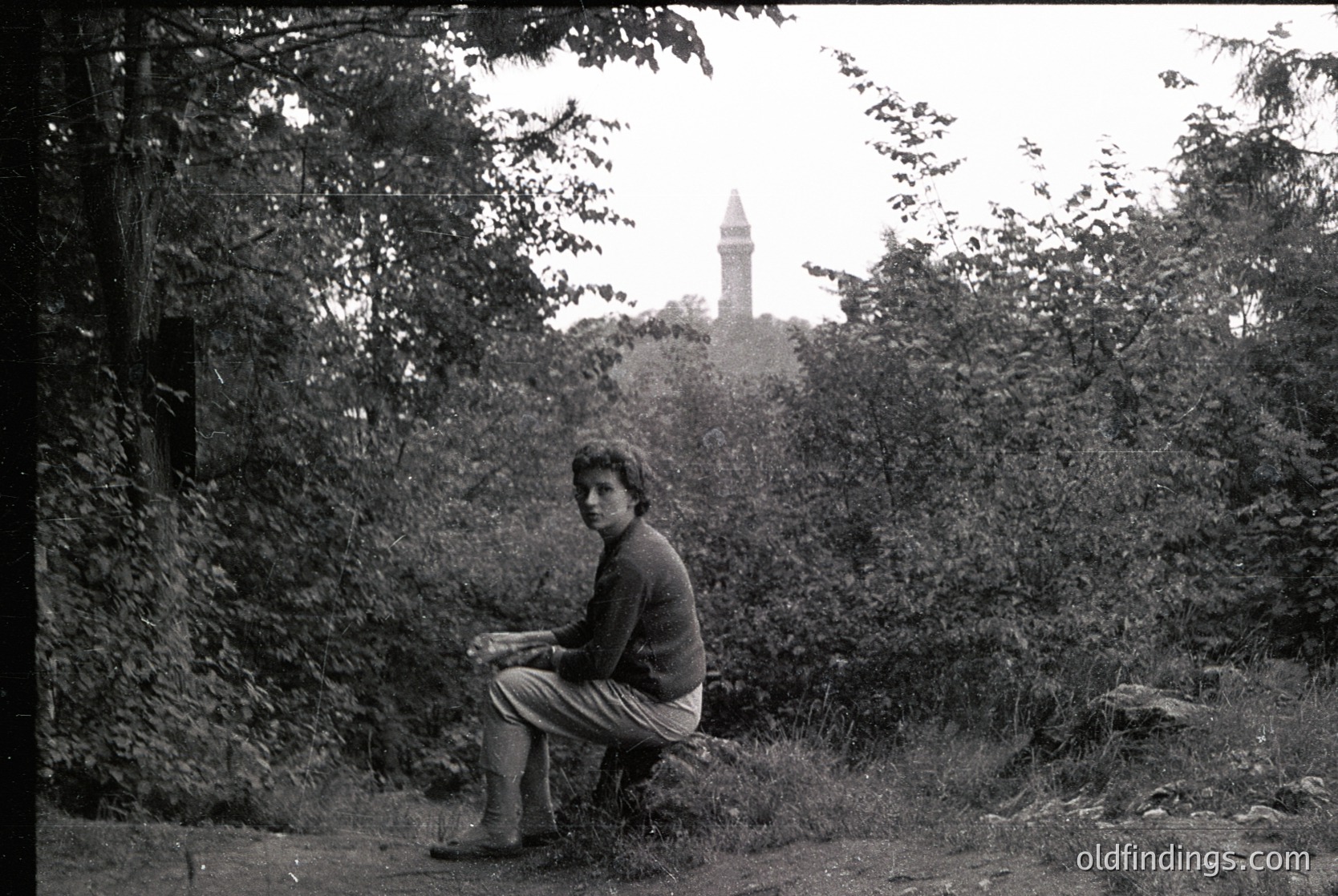 A man sits on a grassy hillside, reading a book, with dense foliage framing the shot. In the background, a tall, cylindrical tower with a conical roof stands amid trees. Likely a 1960s–1970s black-and-white photograph.