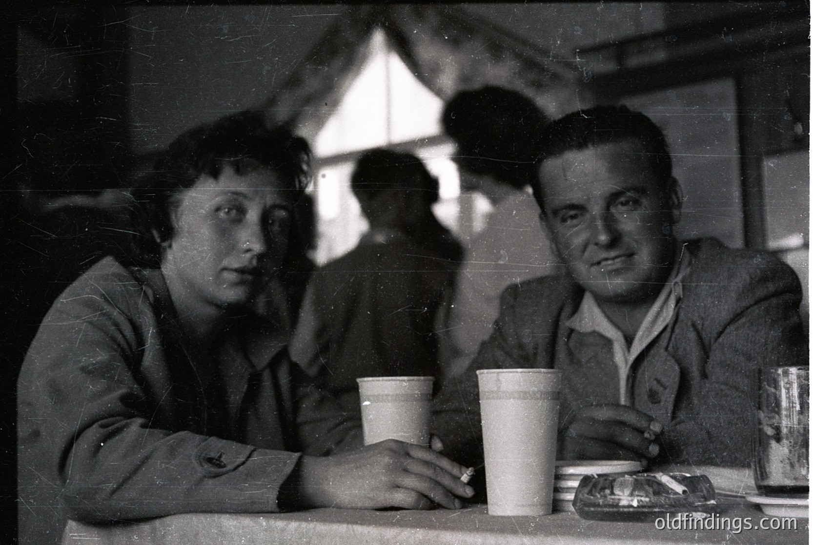 Vintage black-and-white photo of two seated individuals at a café, holding disposable paper cups. Man wears a jacket with a patch, woman in a long-sleeve top. Ashtray and glassware on the table. Soft focus background suggests mid-20th century setting. éCulture