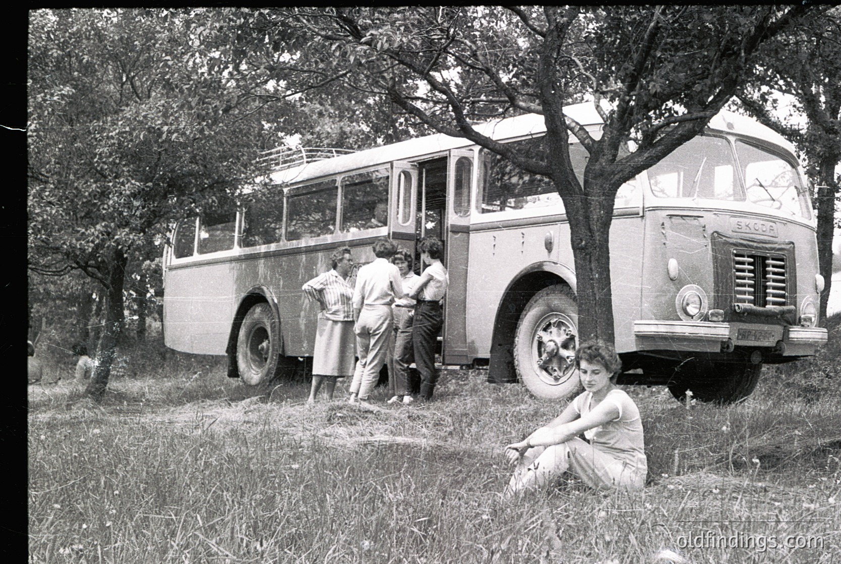 Vintage 1950s-60s scene: group of people near a parked Scania bus in a grassy, wooded area. Four adults and a seated woman in summer attire pose casually. Bus features classic European design with rounded headlights and "Scania" branding.