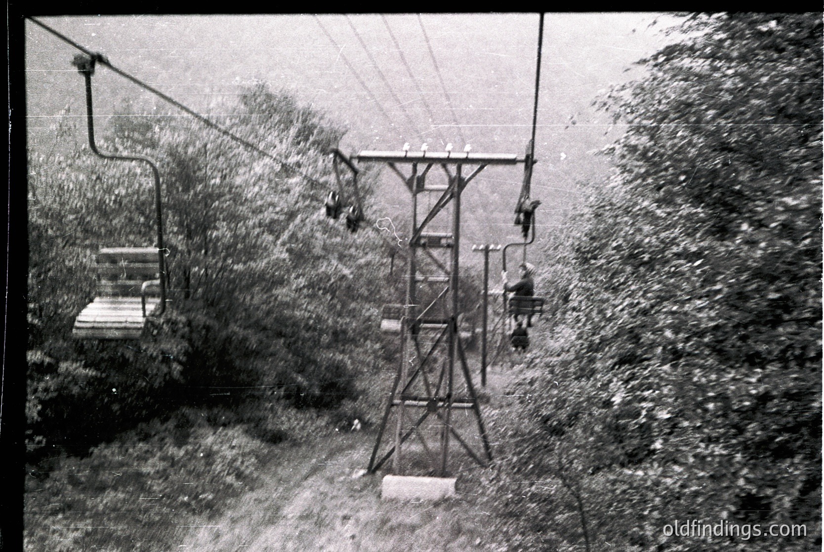 Mid-20th century ski lift with vintage metal chairs suspended on cables, surrounded by dense forest. Snow-covered terrain and heavy foliage suggest winter operations in a mountainous region. Likely or alpine resort infrastructure.