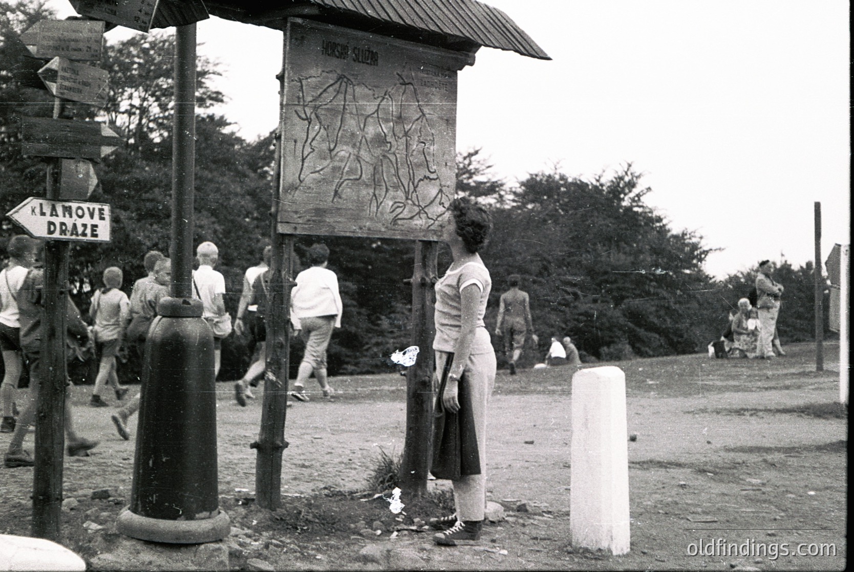 Black-and-white photo of a rural roadside scene with a wooden signpost displaying Cyrillic text ("НАРОДНА СЛОБОДА") and a carved relief of a horse. A woman in a white dress stands near the sign, while others walk or stand around. Signpost also directs to "LANOVE DRAZE." Mid-20th century Eastern European countryside.