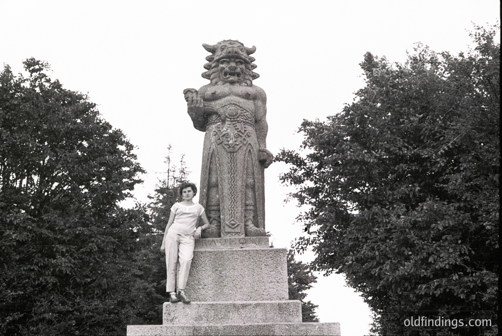Monumental stone statue of a mythical guardian figure with bull’s head and ornate armor, holding a raised sword. Woman in 1960s-style dress poses beside it in a park setting. Likely East Asian cultural or historical site.