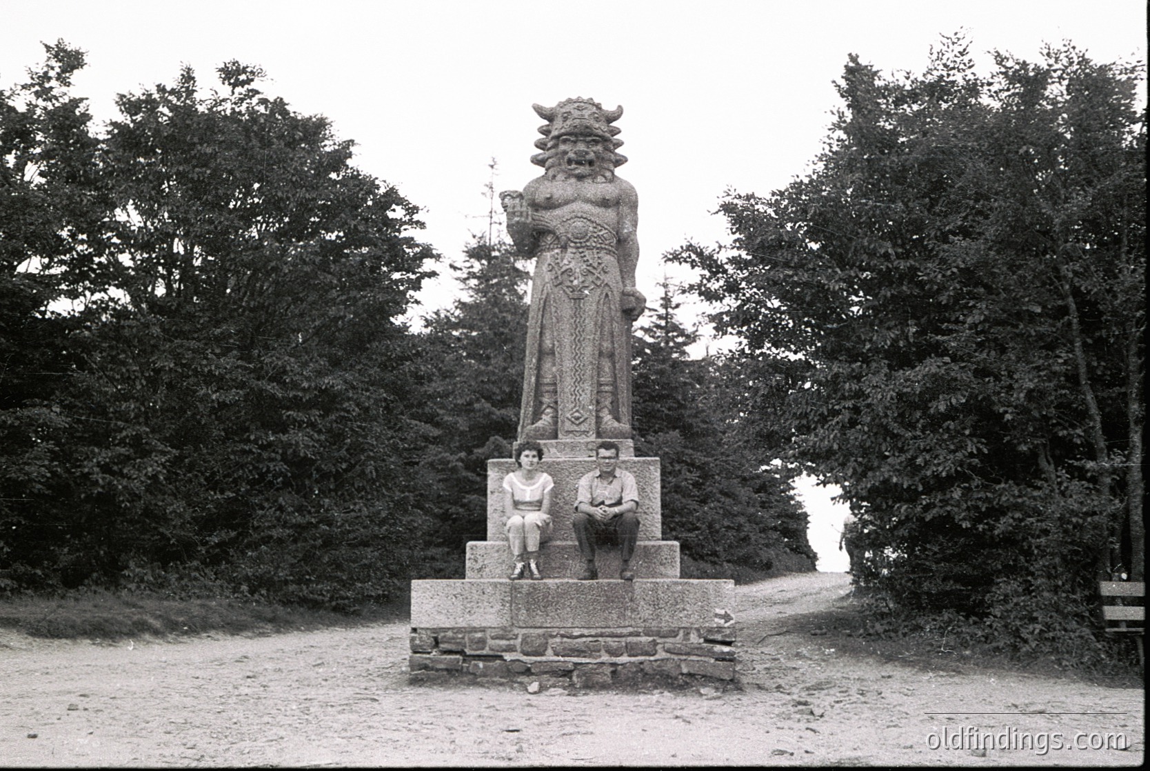Monumental stone statue of a roaring lion-man hybrid atop a pedestal, flanked by two seated figures in 1930s attire. Dense forest and winding path in background. Likely European, mid-20th century.