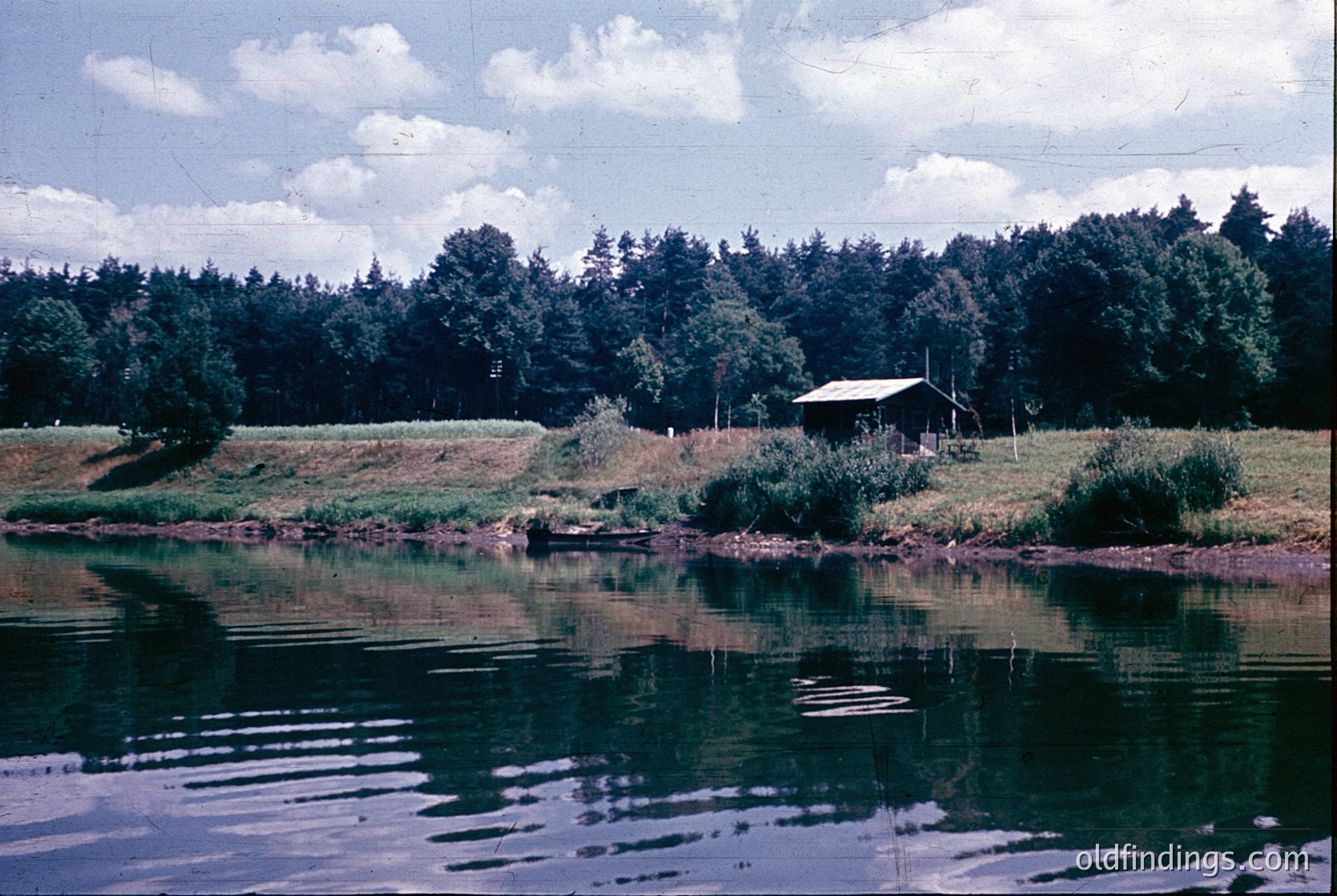 Rustic lakeside cabin nestled in dense forest, reflected in calm waters. Mid-20th century rural setting with wooden structure, overgrown vegetation, and power lines.