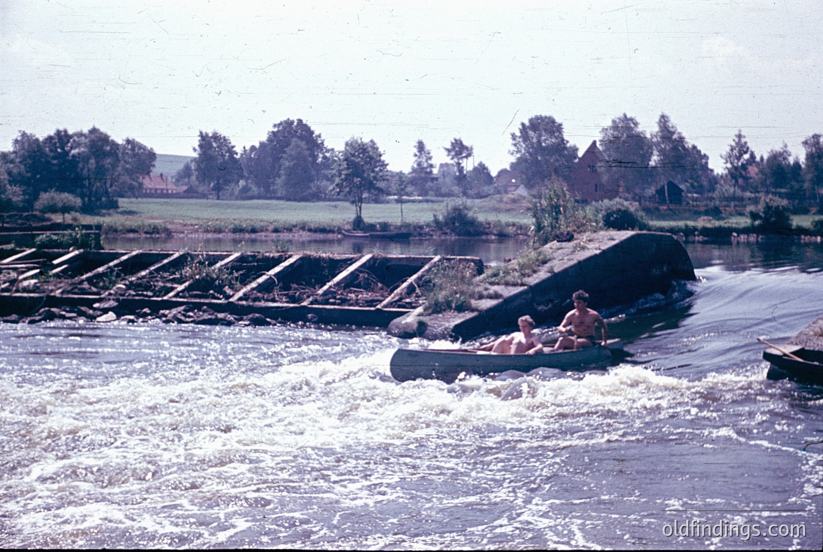 Vintage photo of two individuals in a small wooden boat navigating a rapid on a river, with a concrete dam spillway in the background. Rural landscape features trees, fields, and distant farmhouses. Likely mid-20th century ().