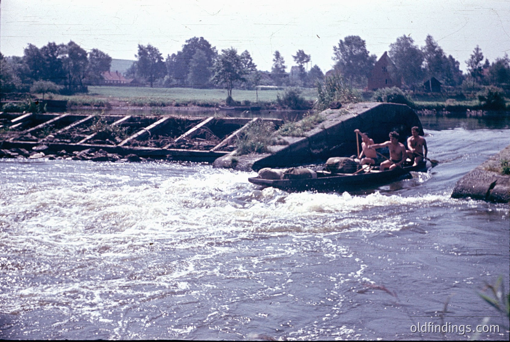 Vintage photo of three individuals rafting on a rapid, using a makeshift raft of logs and tires. Concrete weir with stepped spillway in background. Lush greenery and farmland visible. Likely mid-20th century outdoor recreation scene.