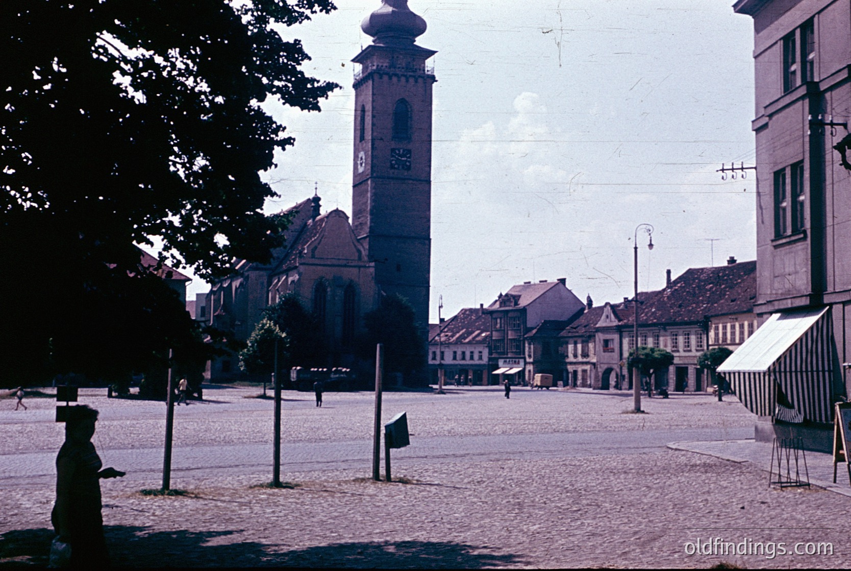 Vintage sepia-toned photo of a European town square, dominated by a tall clock tower with ornate details and a red-tiled church roof. Cobblestone streets and low-rise buildings with gabled roofs frame the scene. A lone figure in dark clothing walks near the foreground, while a striped market stall occupies the right edge. Likely 1950s–1960s, Eastern European architectural style.
