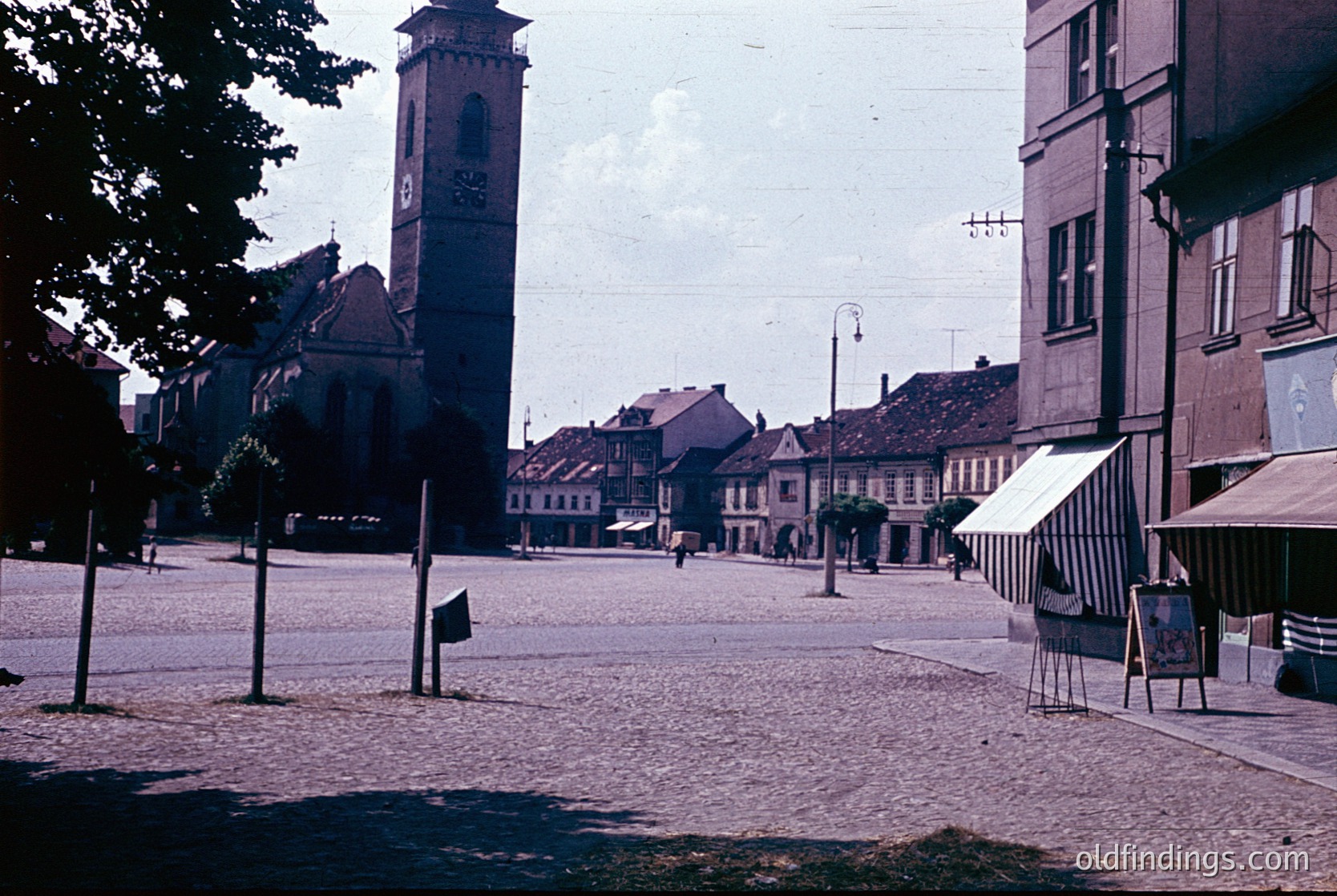 Vintage sepia-toned street scene featuring a prominent clock tower with ornate detailing, likely part of a historic church or town hall. Cobblestone plaza with sparse pedestrian activity, lined by low-rise brick buildings. Striped market canopies hint at local commerce. Overhead wires and vintage signage suggest mid-20th century European setting.