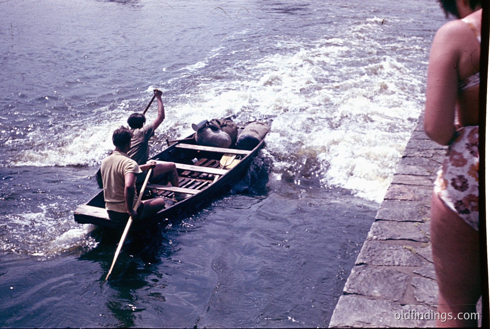 Vintage wooden boat being paddled near a stone pier, mid-20th century. Two men in casual summer attire row while a woman in a floral swimsuit observes from the pier. Waves ripple around the boat, suggesting shallow waters.