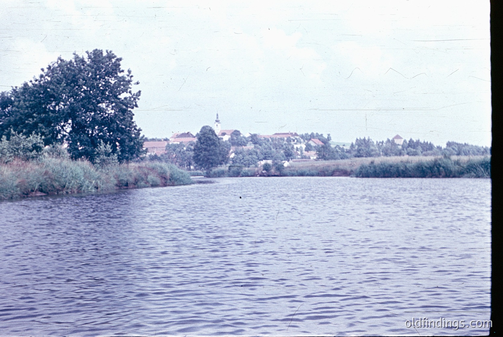 Vintage black-and-white photo of a serene lakeside scene with a church steeple in the background, surrounded by low-lying buildings and greenery. The water’s edge features reeds and a lone tree. Likely Eastern European, mid-20th century.