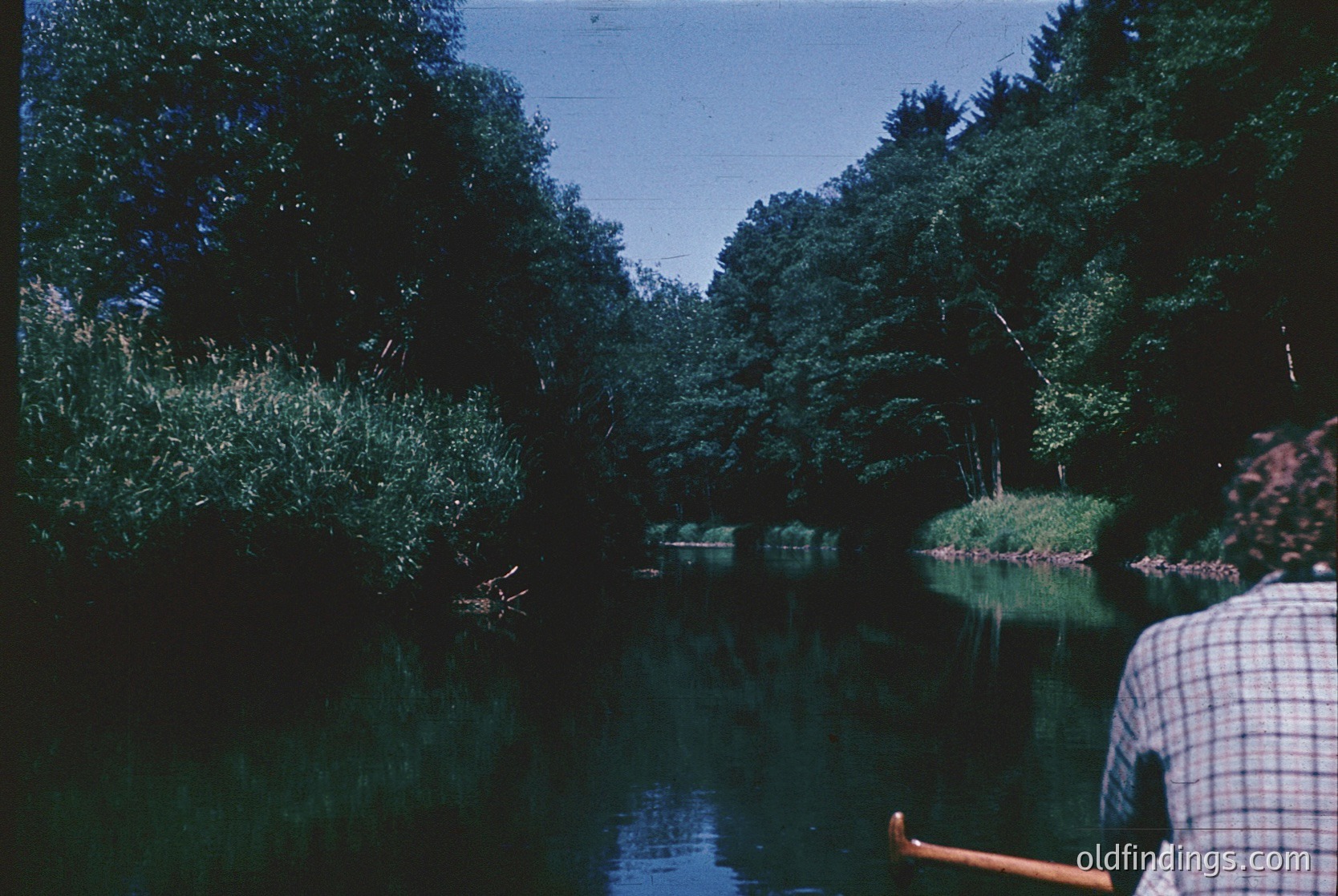 Vintage black-and-white boat on a serene river flanked by dense foliage, suggesting mid-20th century recreational boating. Overcast skies enhance the moody, reflective water surface. Partial view of a person in a checkered shirt paddling, indicating leisure activity.