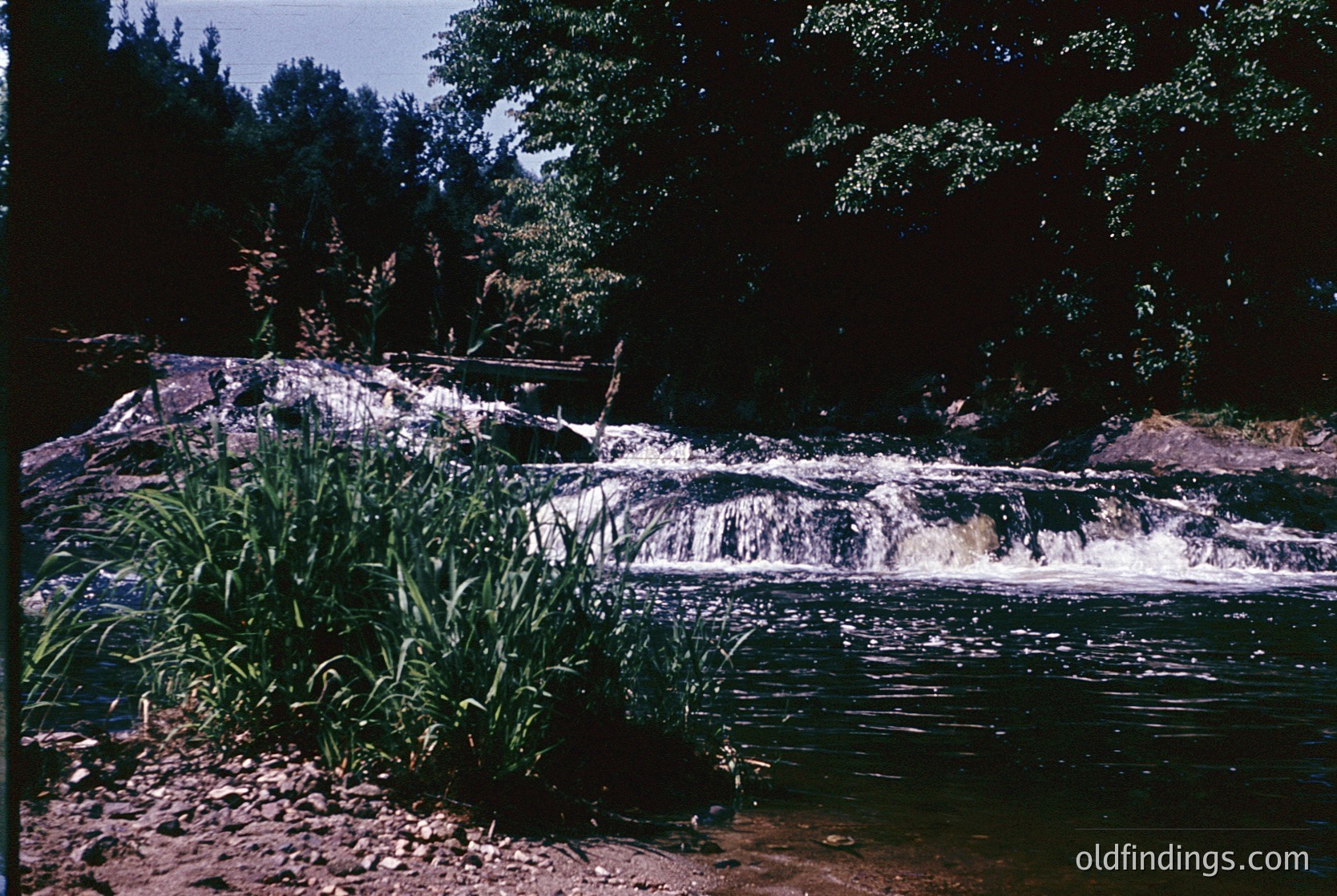 Vibrant 1970s-era waterfall cascading over layered rock formations into a calm pool, surrounded by dense foliage. Warm sepia-toned filter enhances natural hues. Ideal for nature, landscape, and environmental studies.