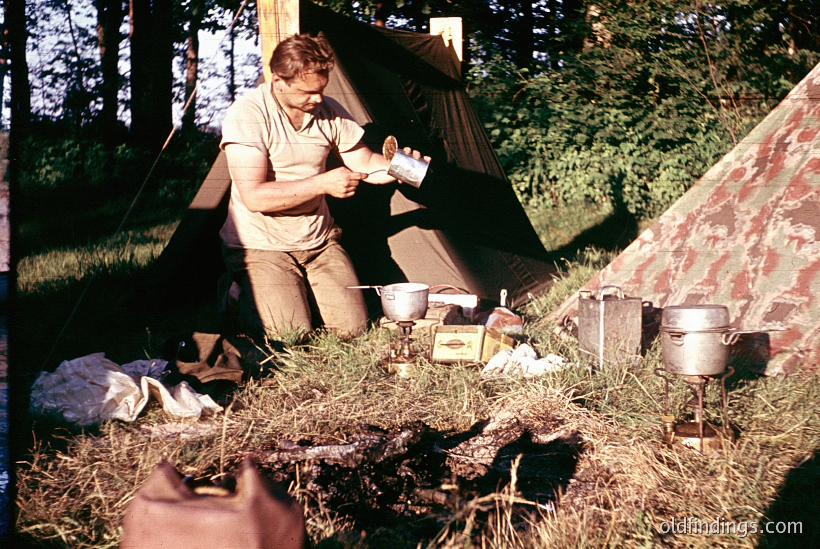 Man preparing food at a campsite in a forested area, 1960s–70s. Tent, vintage camping gear (metal pots, canned goods, cloth bags) and hay-covered ground visible. Classic outdoor lifestyle.