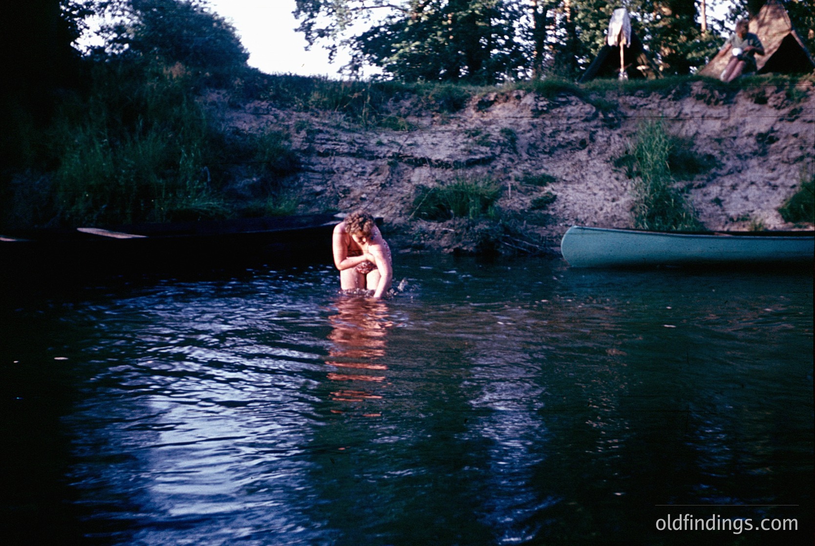 Vintage 1960s/70s outdoor scene: Couple embracing in shallow river, surrounded by lush greenery and rocky banks. Green canoe rests on right. Warm sepia tones enhance nostalgic feel.