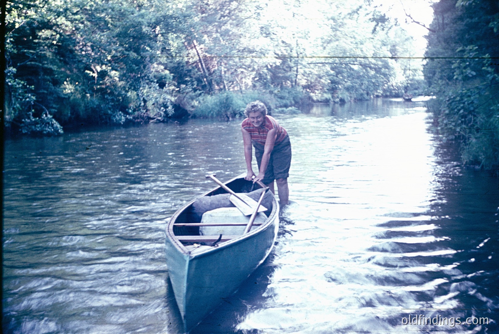 Vintage canoeing scene: man in 1970s-style plaid shirt and green pants prepares to launch a sleek, narrow canoe on a calm river surrounded by dense foliage.