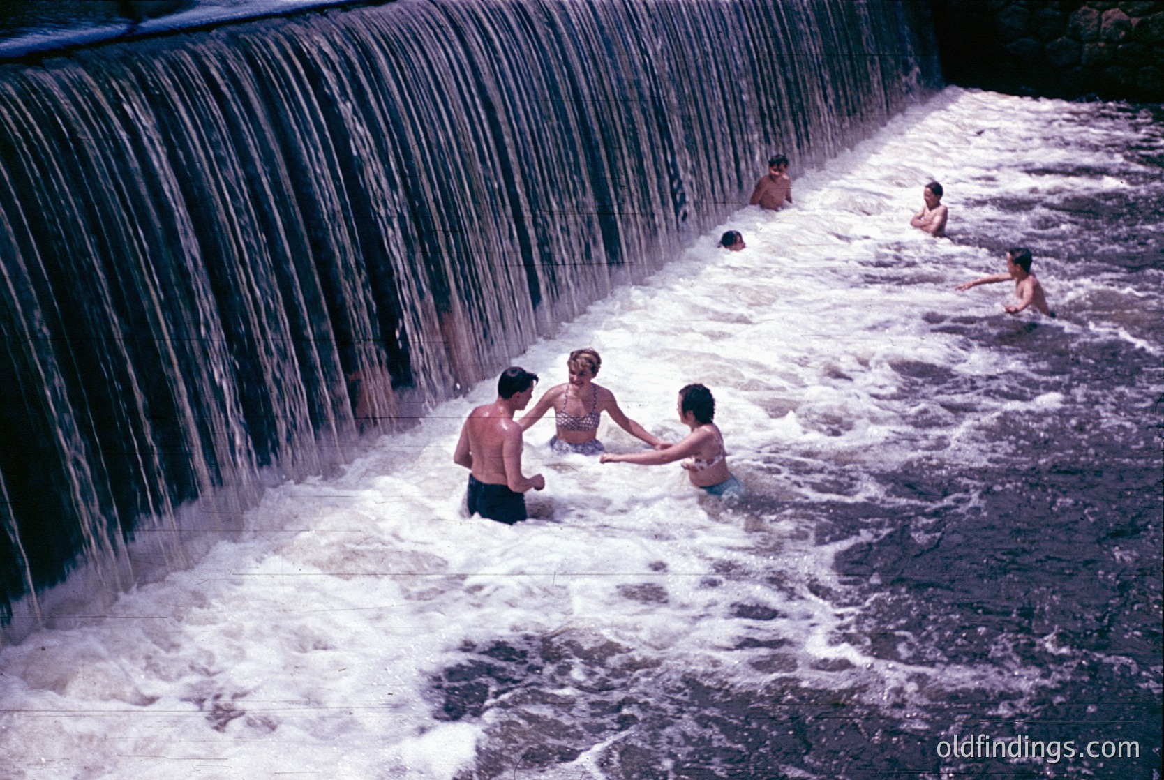 Vibrant 1960s/70s scene of children playing beneath a cascading waterfall. Concrete dam creates a natural pool; three kids in foreground hugging, others swimming. Warm sepia tone enhances nostalgic feel. Ideal for retro lifestyle or family bonding references.