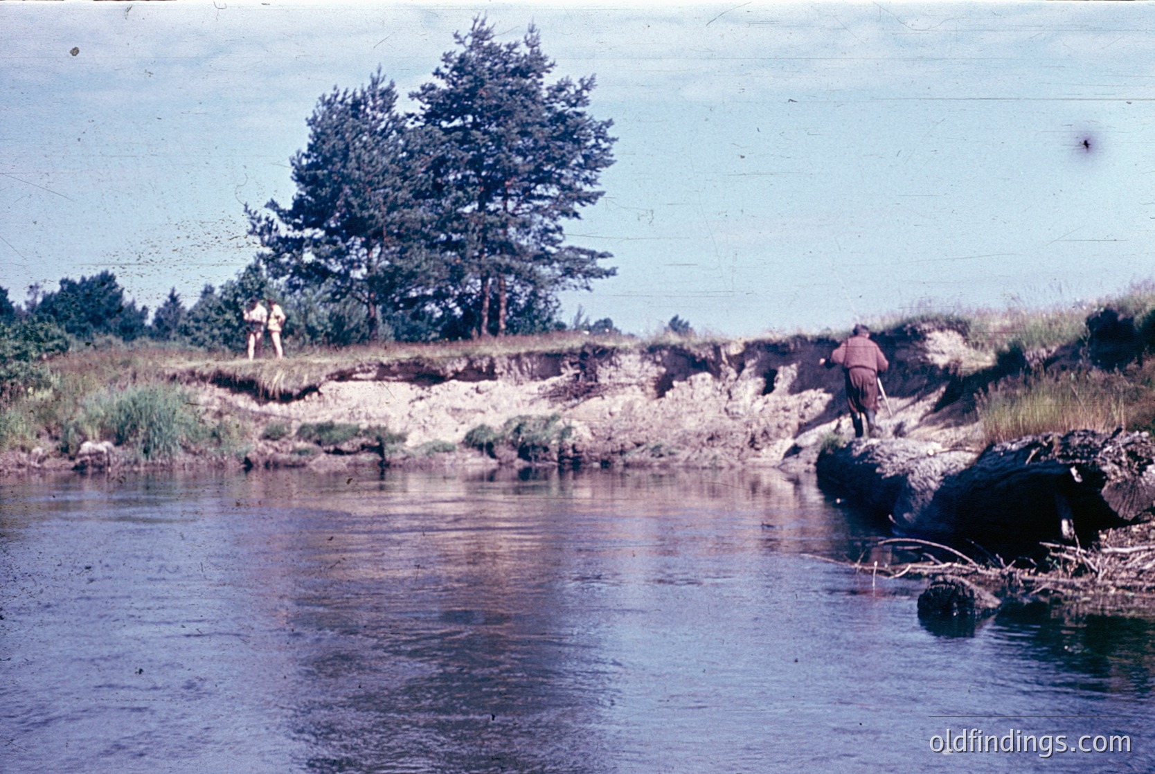Vintage black-and-white photo of two individuals wading in a shallow riverbed surrounded by rocky terrain and sparse vegetation. The person on the right wears a long-sleeve shirt and shorts, while the other stands near a lone tree. Likely mid-20th century, rural setting.