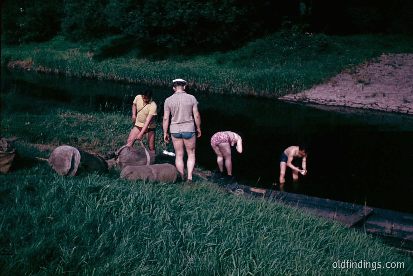 Four individuals in 1960s-style swimwear gather near a shallow, terraced waterway, likely for a communal dip or water activity. The man in a light-colored cap and shorts stands near a large rock, while three women—one in a yellow top, another in a pink swimsuit, and a third in a light dress—lean or kneel by the water’s edge. Lush greenery and terraced fields frame the scene, suggesting a rural or agricultural setting. The sepia-toned filter enhances vintage appeal.