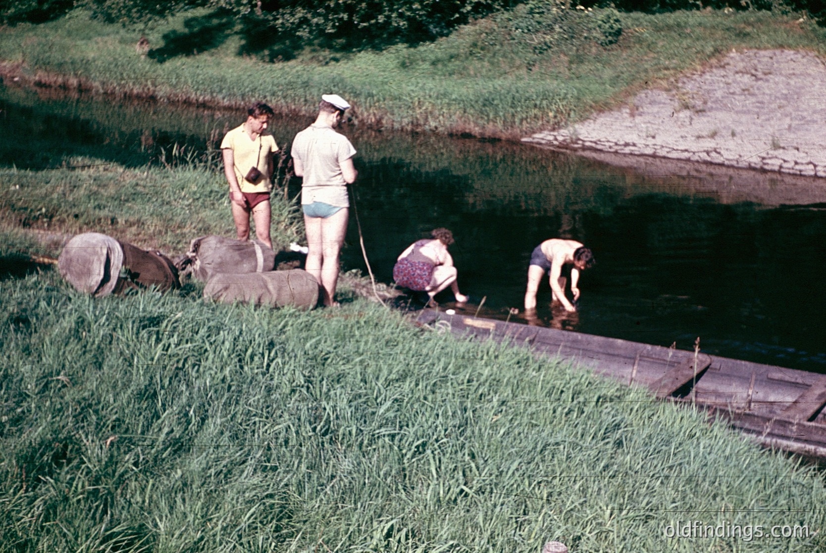 Four individuals in 1960s-style swimwear repair a wooden boat on a grassy riverside bank. The scene captures rural labor, likely for fishing or transport. Vibrant sepia-toned filter suggests vintage photography.