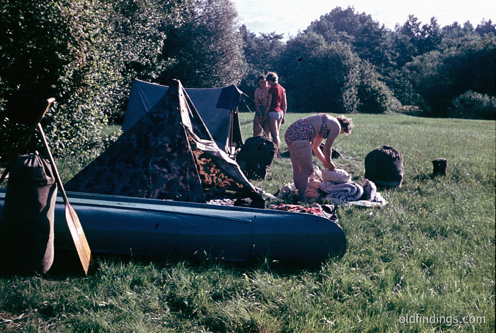 Vintage outdoor camping scene with inflatable raft and vintage tent. Two adults (one in floral dress, one in striped shirt) set up gear in a grassy field under trees. Inflatable raft with paddles and picnic blanket with bags nearby. Likely late 20th century, midwestern U.S. or Europe.