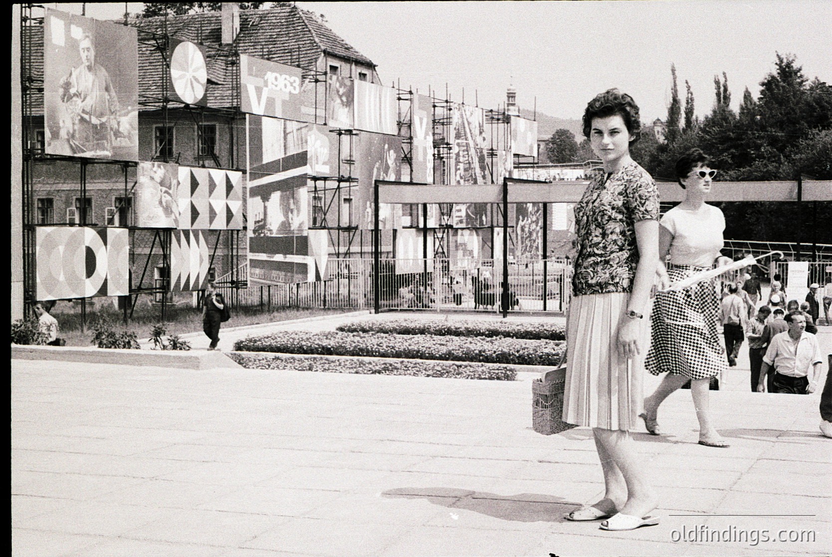 Mid-century modern courtyard with geometric murals and "1968" date plaque. Two women in 1960s fashion—one in a floral blouse and pleated skirt, the other in a sleeveless dress—walk near a decorative fountain. Concrete architecture and urban greenery blend in this public space.
