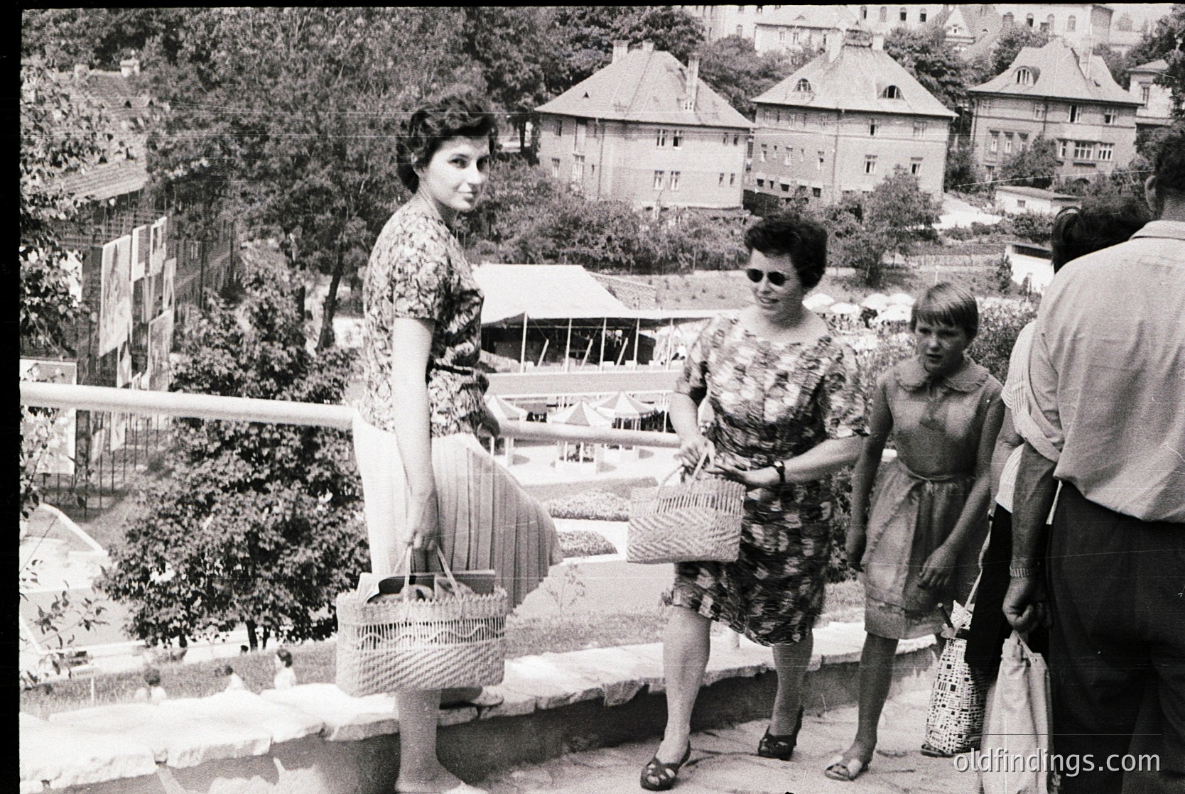 Black-and-white street market scene featuring three women in 1960s Eastern European attire—floral dresses, sunglasses, and woven baskets. Stalls display textiles and household goods under awnings. Residential buildings with balconies and greenery in background.