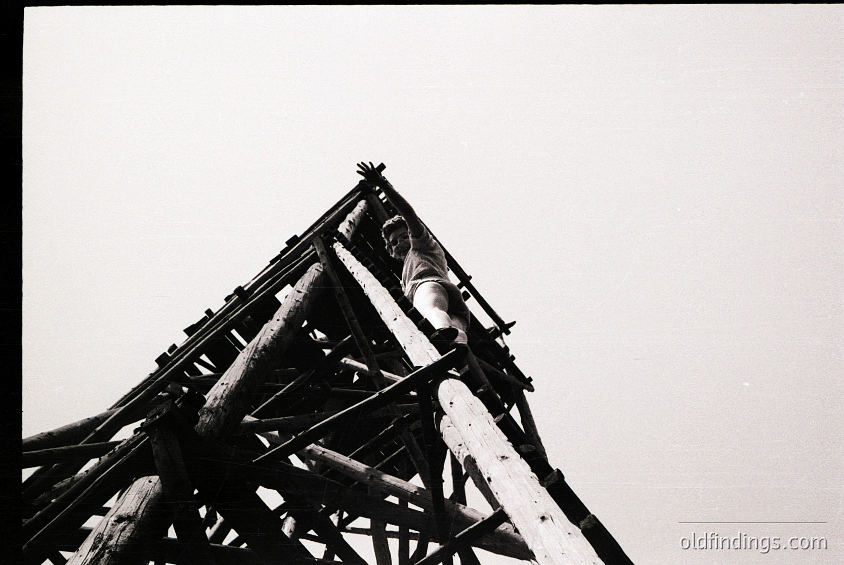 Black-and-white industrial shot of a wooden crane structure with a worker perched on the upper beam, likely mid-20th century. Distinctive lattice framework and exposed beams highlight industrial architecture.