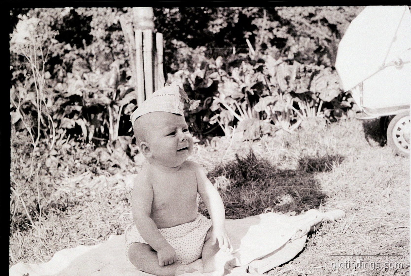 Mid-20th century black-and-white photo of a toddler in outdoor setting, wearing a sailor-style cap and diaper. Lush greenery and a vintage car in background suggest a suburban or rural garden. Candid, nostalgic family moment.