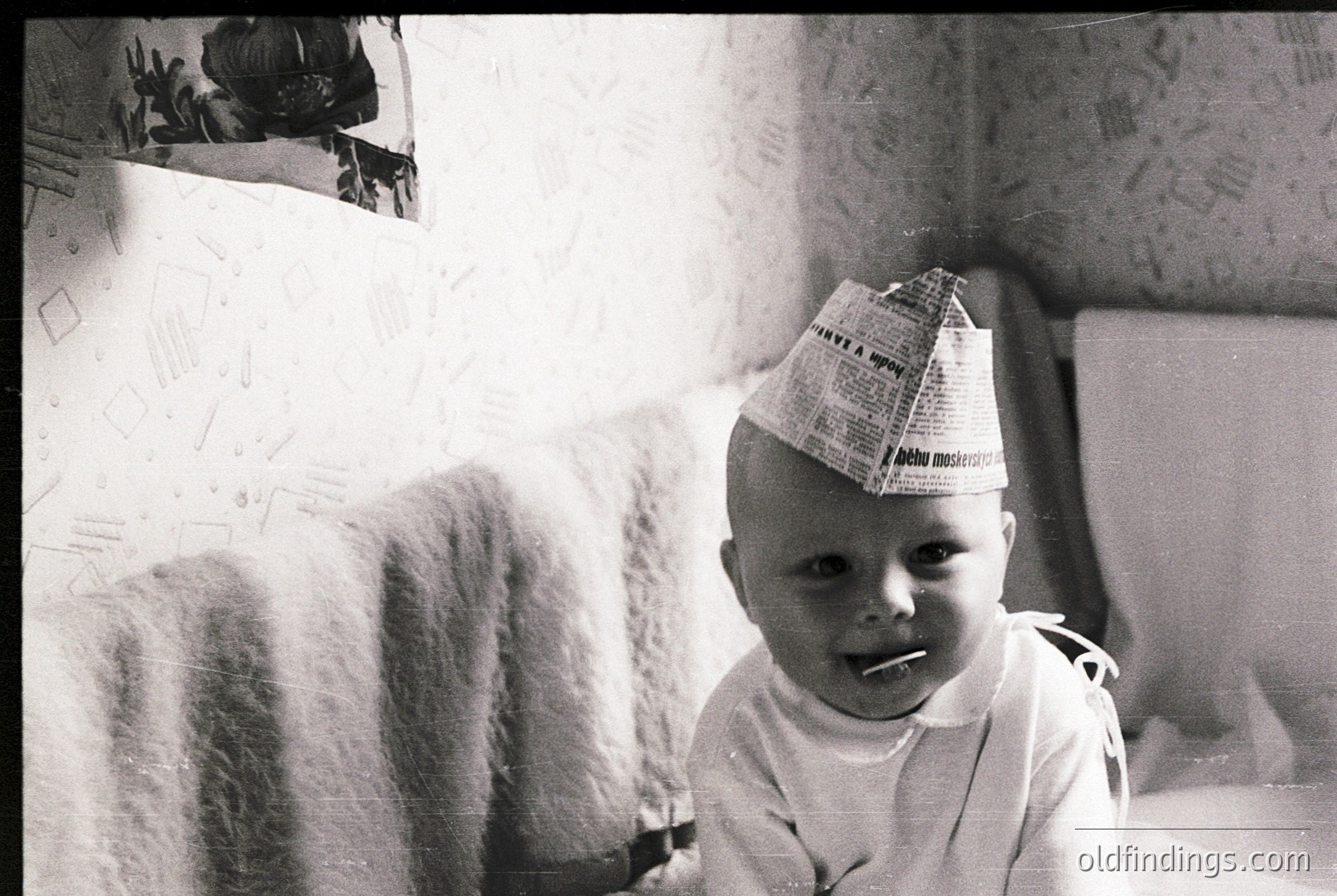 Vintage black-and-white photo of an infant wearing a newspaper hat and light-colored onesie, seated indoors. Wall displays handwritten notes and a framed photo. Mid-20th century domestic setting.