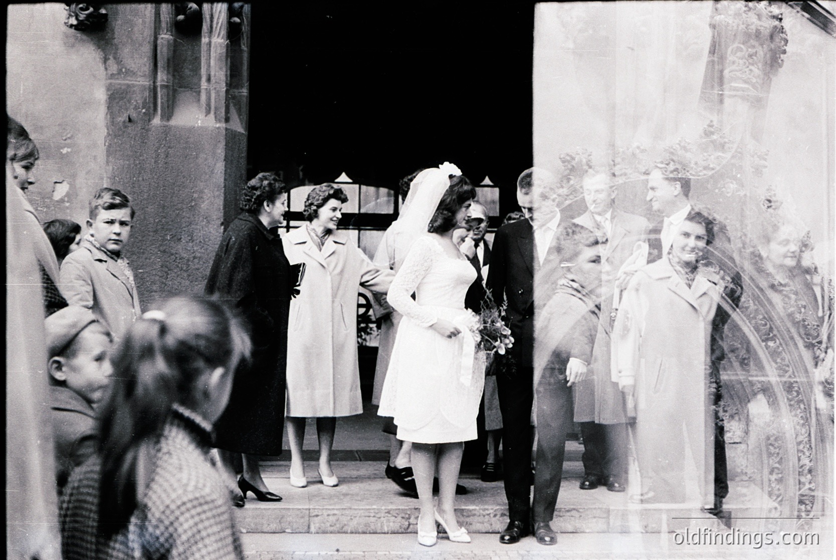 Vintage black-and-white wedding photo featuring a bride in a full-length white gown with a veil, exiting a grand building with ornate stonework. Mid-20th century attire—men in suits, women in floor-length dresses—suggests 1950s–1960s European setting. Crowd of onlookers, including children, captures communal celebration. Architectural details hint at historic urban location.
