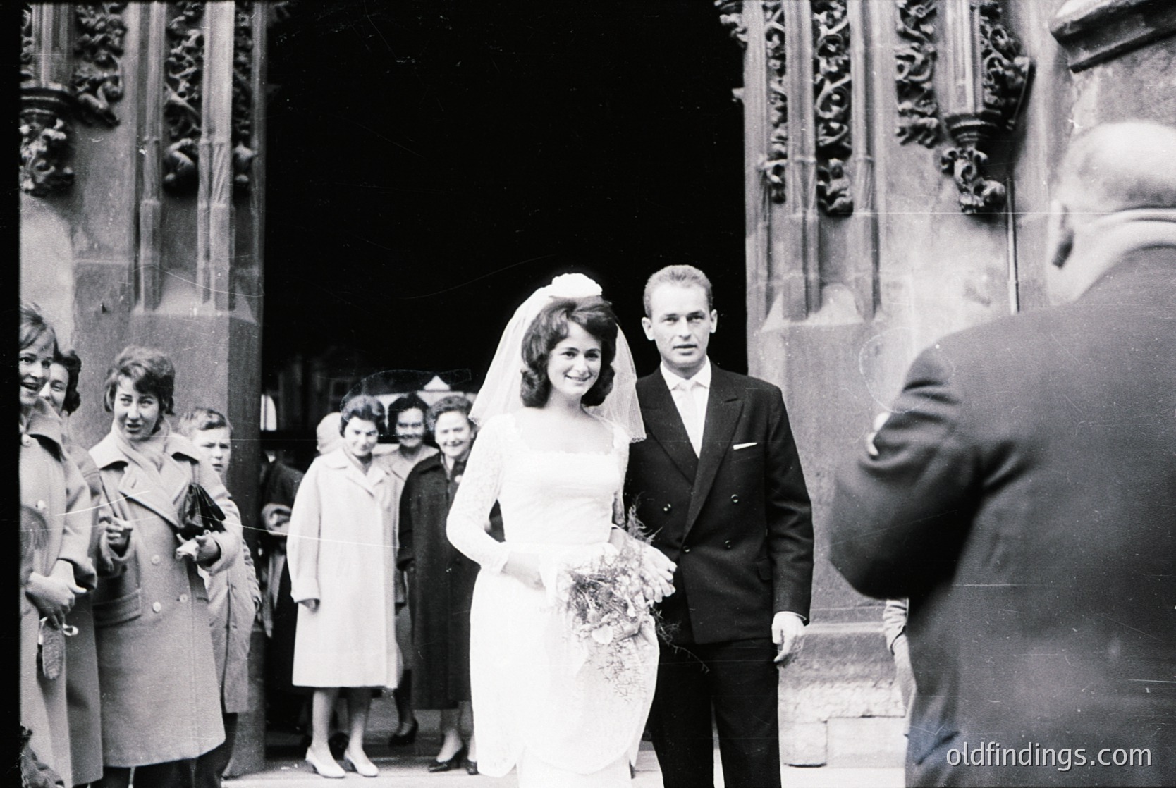 1960s black-and-white wedding photo outside ornate church. Bride in full-length gown with veil, bouquet, and groom in dark suit. Crowd in mid-century attire. Architectural details: carved stone columns, arched doorway.