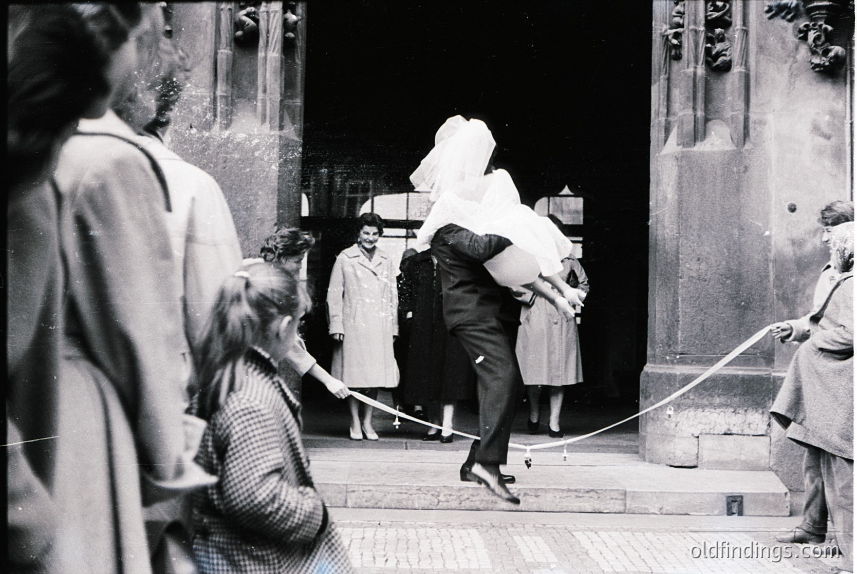 Mid-20th century black-and-white photo: newlywed couple exiting church in Berlin, 1950s. Bride in white veil, groom in suit, surrounded by well-wishers on stone steps. Architectural details include ornate doorway and rope barrier. Ideal for historical research on post-war German weddings.