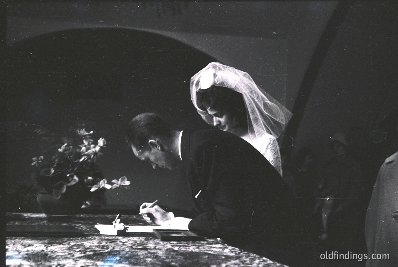 Black-and-white wedding ceremony shot featuring bride and groom signing a register. Bride wears veil and formal gown; groom in suit. Floral arrangement and ornate arch in background. Likely 1950s–1970s Western tradition.