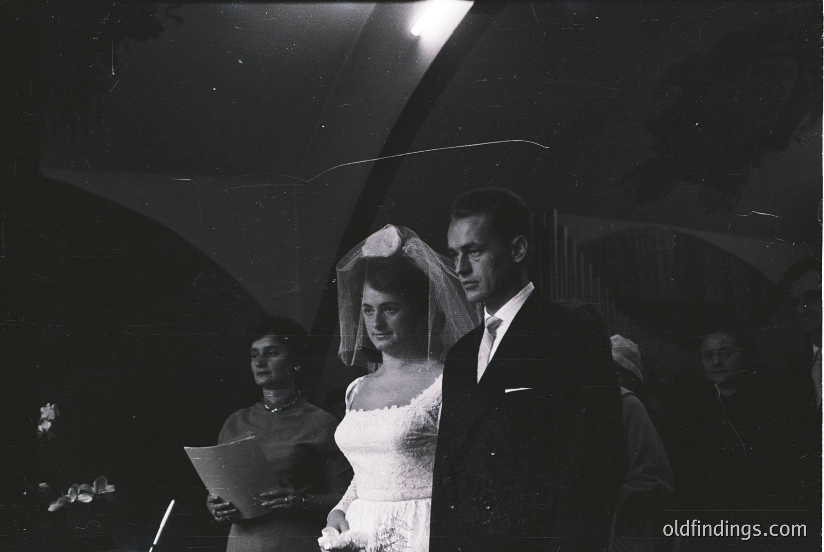 Mid-20th century black-and-white wedding portrait: bride in classic veiled gown, groom in dark suit, holding hands under dramatic stage lighting. Formal ceremony setting with blurred audience and hymnal in focus.