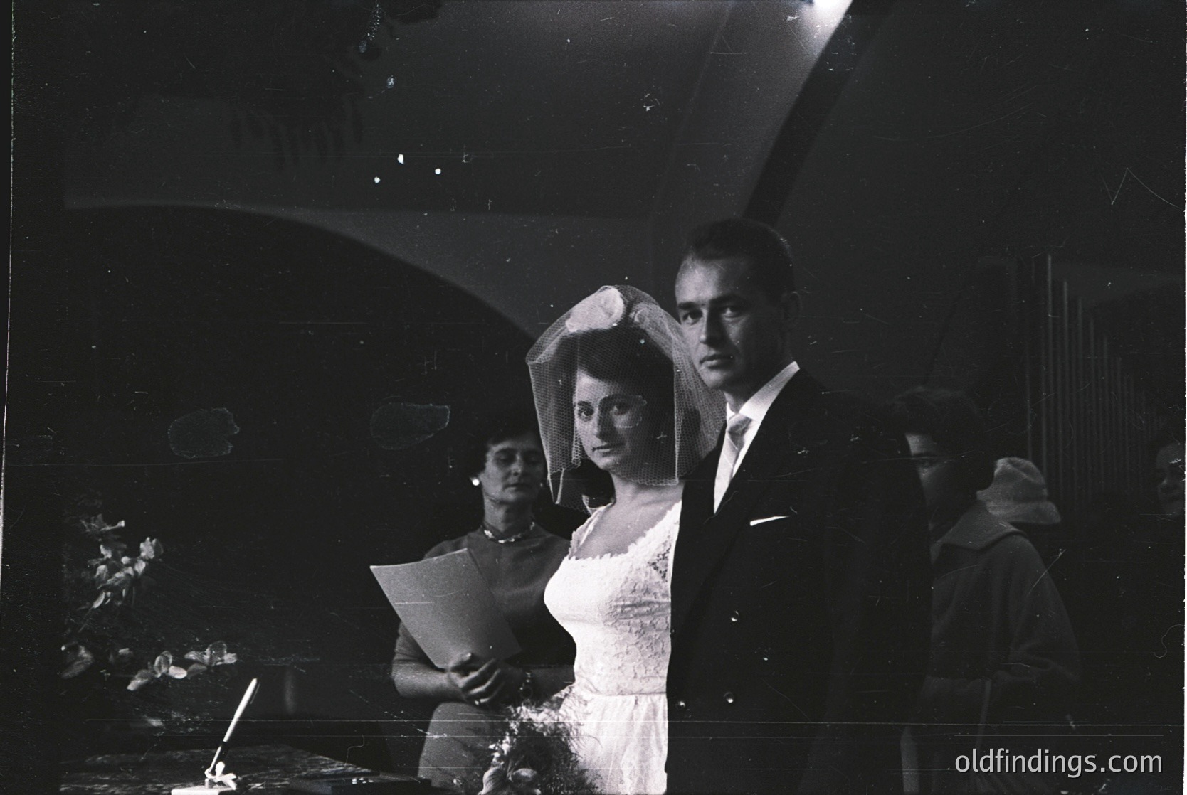 Black-and-white wedding portrait featuring a bride in a full veil and groom in formal attire, likely 1950s–1970s. Indoor setting with blurred guests and a candlelit table. Classic mid-century wedding style.