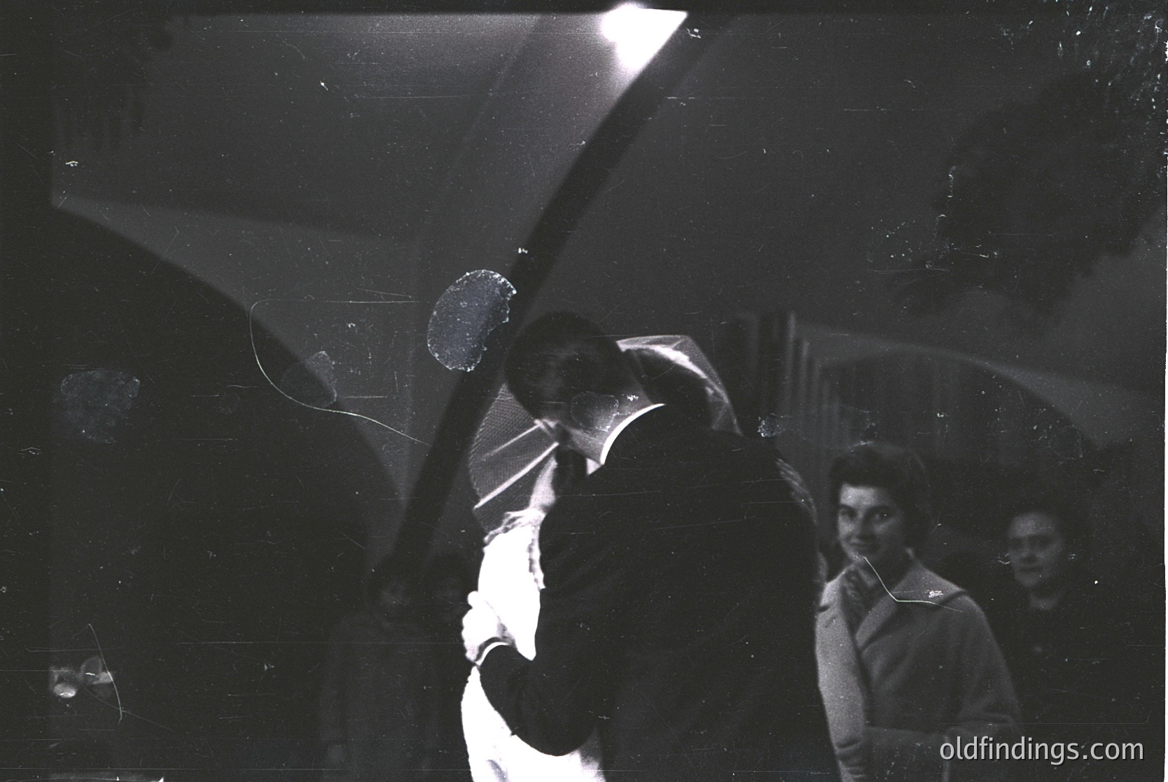 Vintage black-and-white photo of a couple dancing closely in a dimly lit room, likely a 1950s–1960s social event. The man’s arm wraps around the woman’s waist, while a spotlight illuminates them from above. Background shows blurred figures, suggesting a crowded venue. Film grain and lighting effects enhance nostalgic atmosphere.