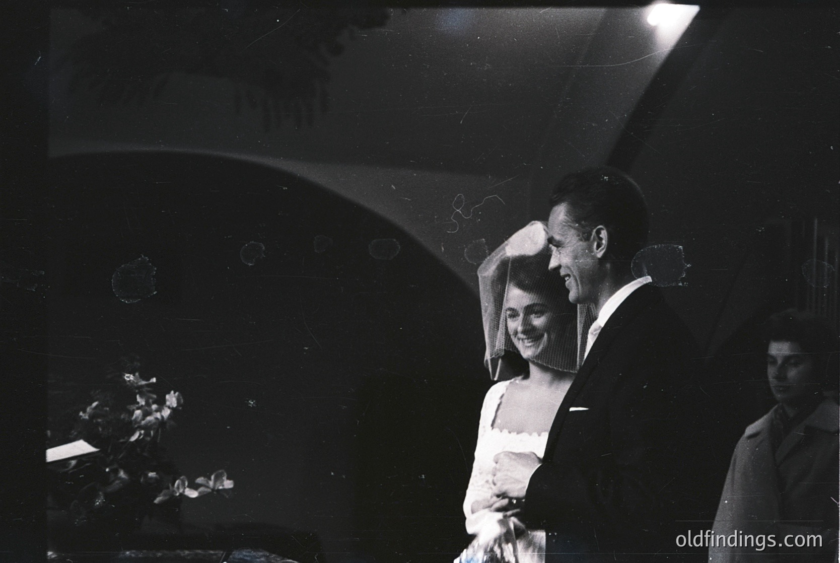 Couple in vintage wedding portrait, 1950s-1960s. Woman in full-length gown with veil, man in dark suit adjusting her veil. Soft spotlight and blurred background suggest formal indoor setting. Candid, joyful moment captured in black-and-white.