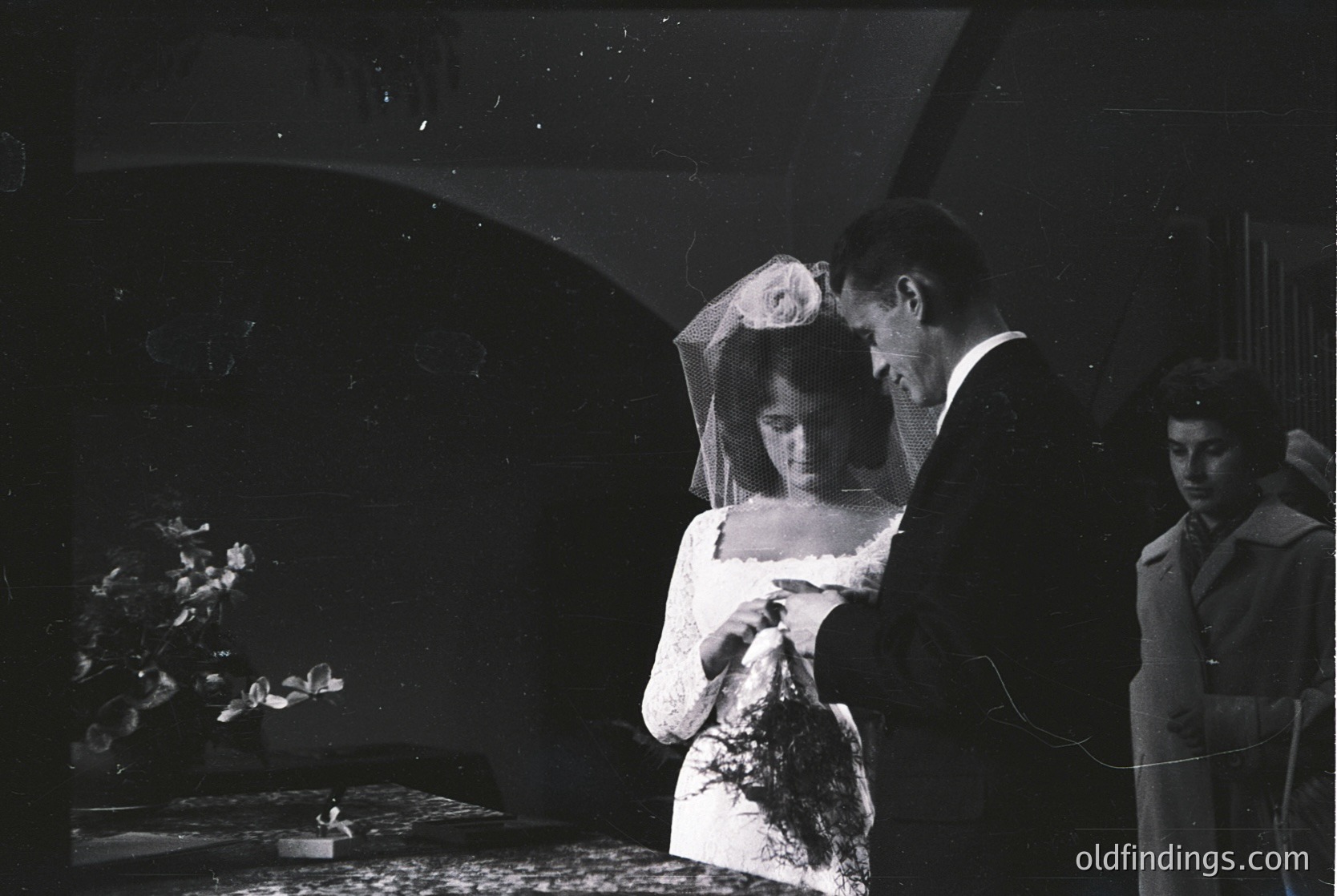 Mid-20th century black-and-white wedding portrait: bride in lace veil and formal gown, groom adjusting her veil, third person (likely photographer) in suit. Indoor setting with floral centerpiece and draped curtains.
