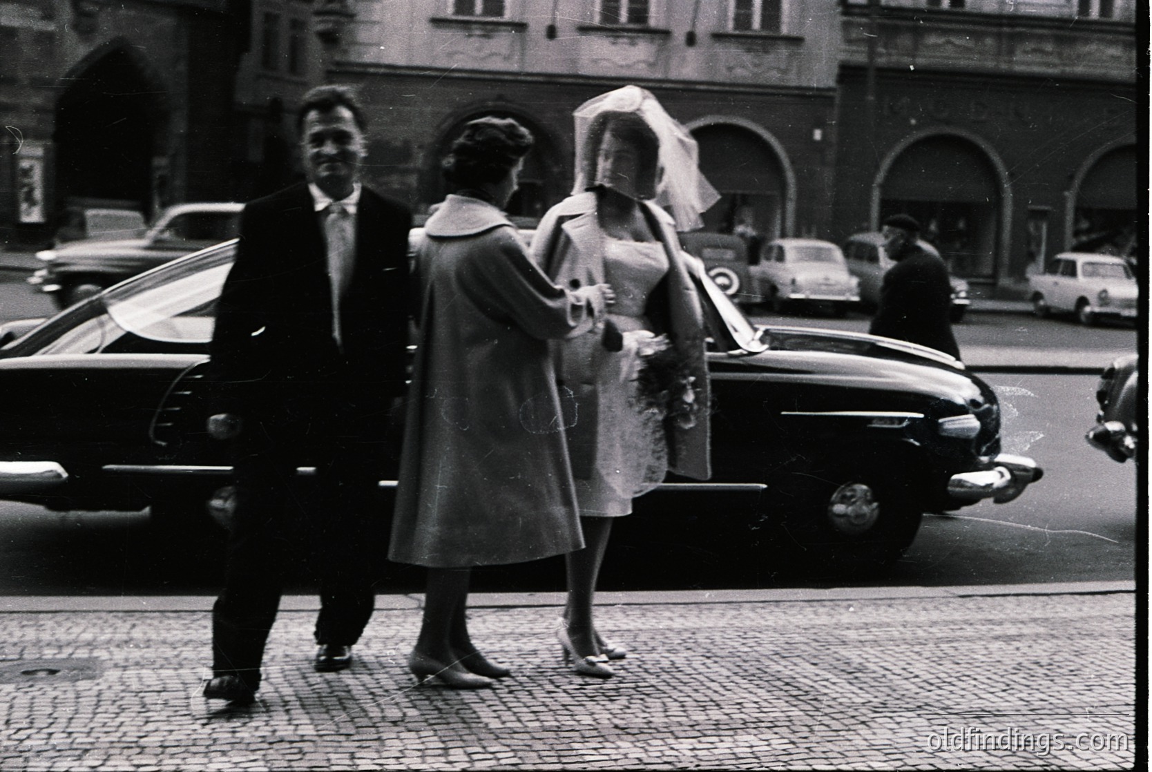 Classic 1960s black-and-white street scene: newlywed couple exiting vintage car (likely Jaguar XK120) in urban setting. Bride in full veil, groom in suit; matron of honor in coat. Cobblestone road, mid-century architecture, and parked cars suggest European city vibe.