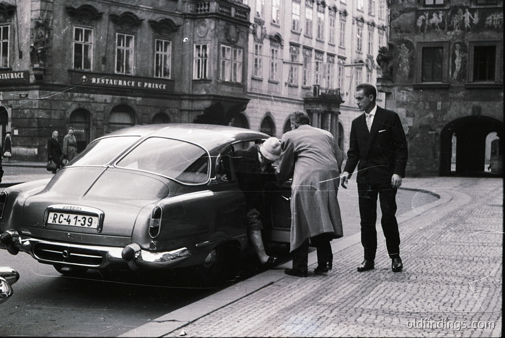 Classic 1950s-60s European street scene featuring a **Mercedes-Benz 300SL "Gullwing"** (RC-41-139 license) parked on cobblestone. Two men in suits and a woman in a coat assist a child exiting. Historic **Prague** architecture with ornate facades and "Restaurant U Prince" signage visible. Black-and-white vintage style captures mid-century urban life.