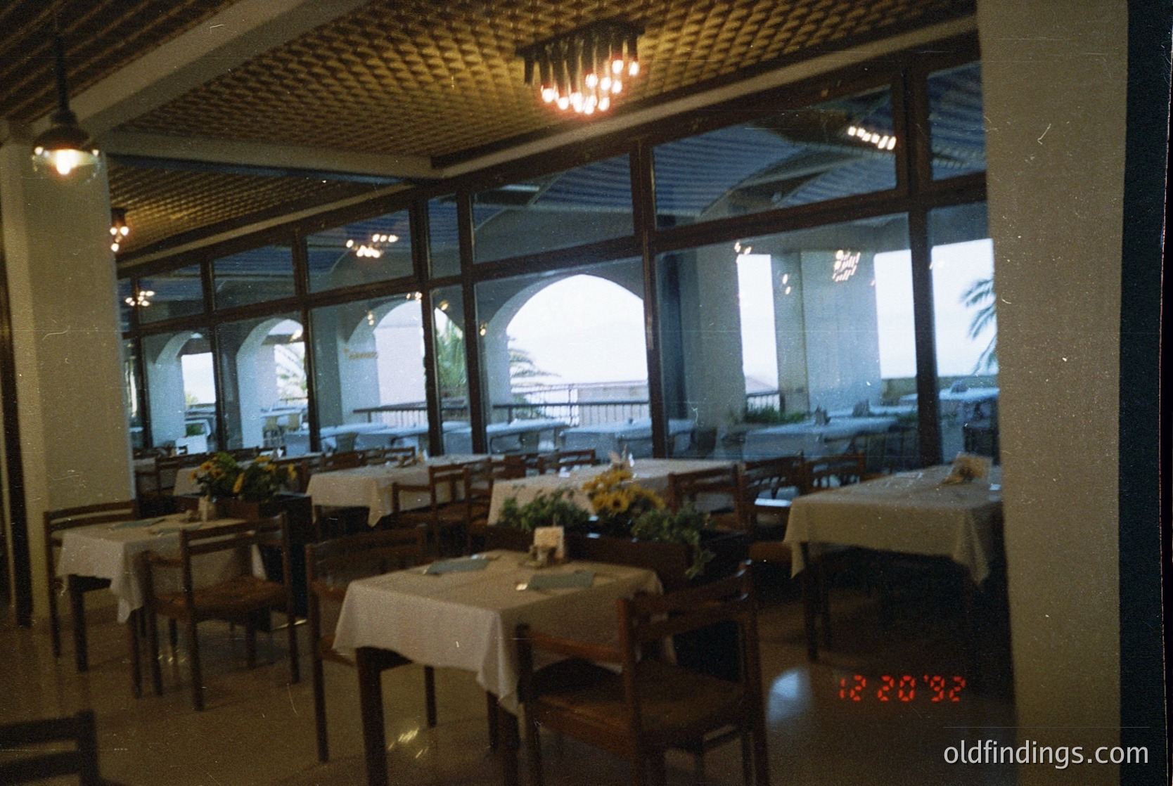 Mid-century seaside restaurant with arched windows overlooking ocean. Wooden tables draped in white linen, centerpiece flowers, and vintage lighting. Date stamp suggests . Architectural style blends modernist with coastal influences.