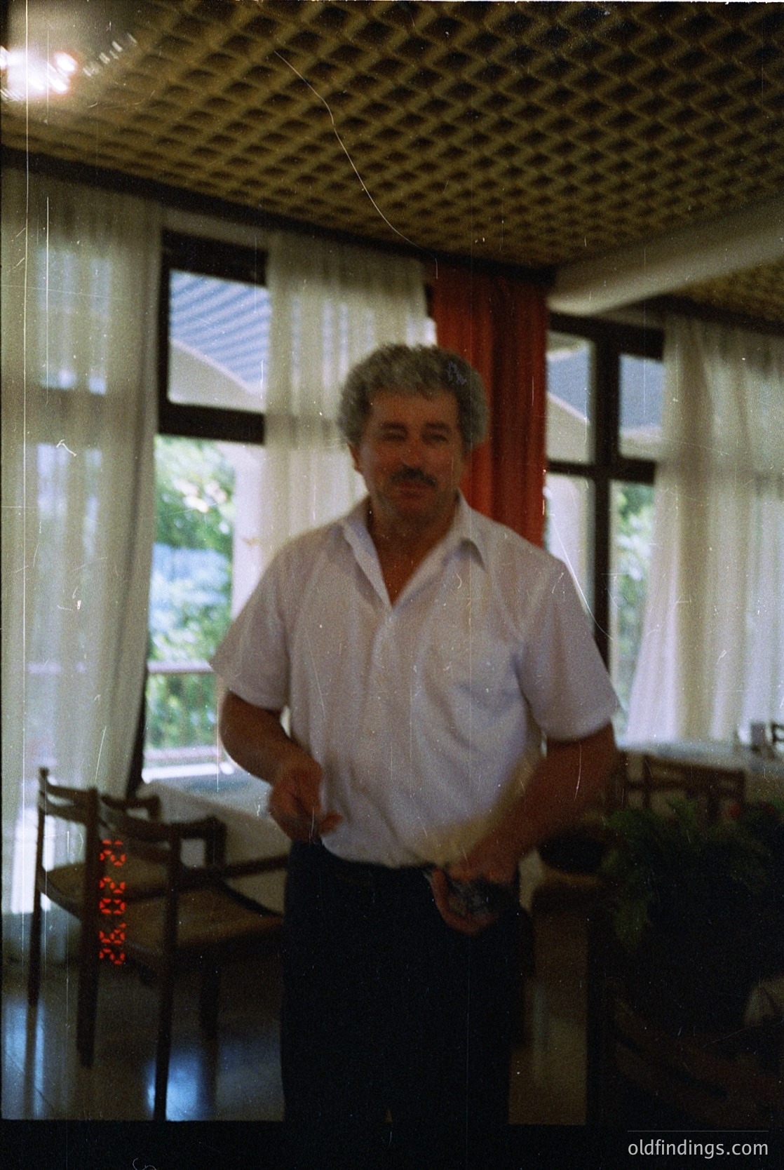 Mid-century indoor portrait of a man in a light button-down shirt and dark trousers, standing beside a wooden table with a digital clock displaying "10:35." Checkered ceiling and sheer curtains framing a view of greenery outside. Likely 1970s–1980s residential or office setting.