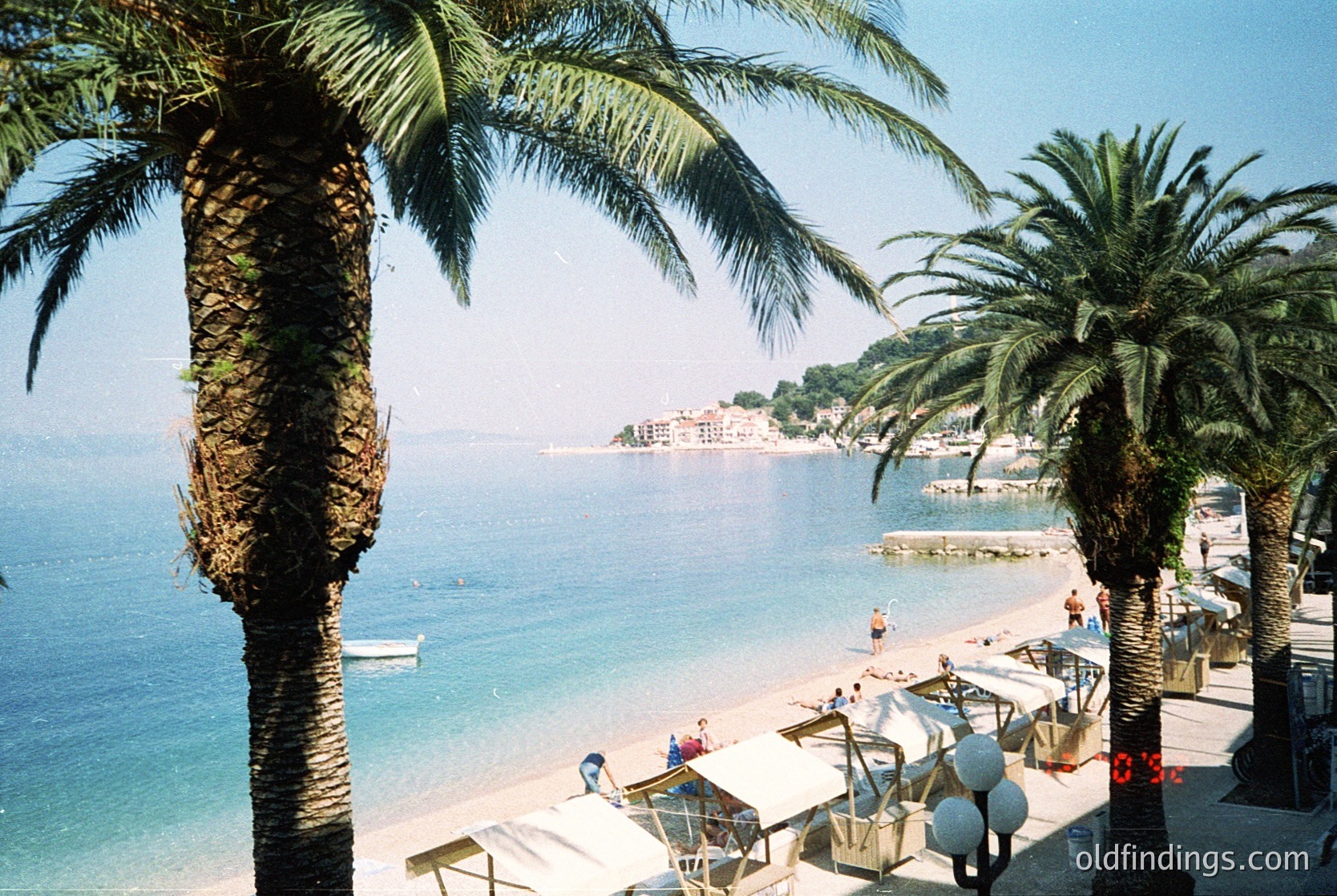 Vibrant seaside promenade lined with tall palm trees and beach umbrellas. Clear blue waters meet a rocky coastline with scattered buildings. Lifeguard tower and beachgoers visible. Likely Mediterranean coastal town, mid-20th century.