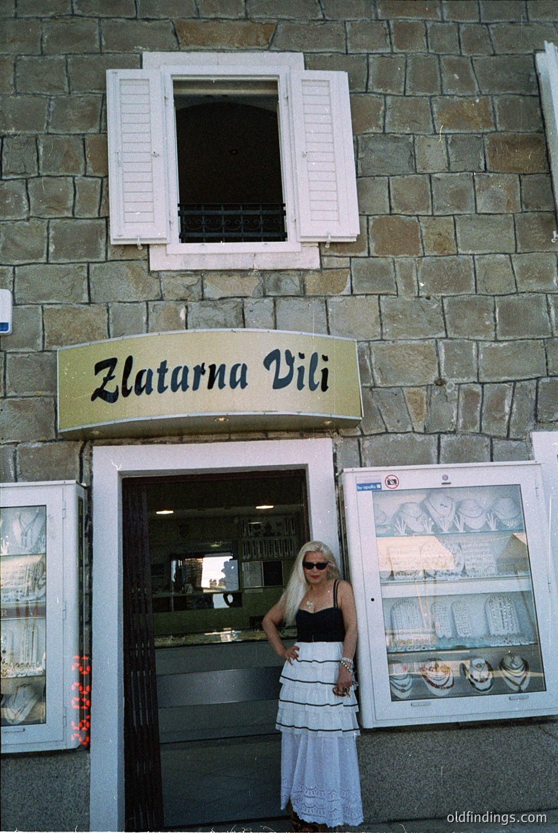 Vintage gold sign "Zlatarna Vili" above a stone-framed shop entrance, showcasing jewelry displays inside. Woman in 1970s-style dress poses near open door. Stone facade with white shutters and framed posters.